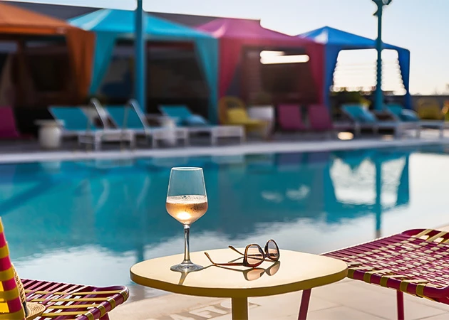 A wine glass and sunglasses on a small table beside a pool with colorful canopies and lounge chairs in the background.