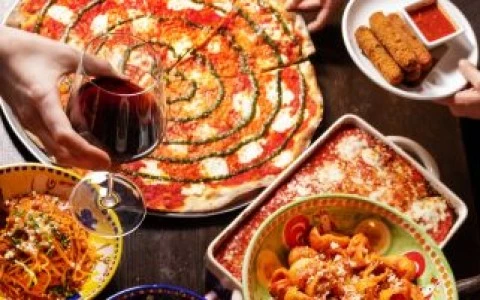 A table with a variety of food including pizza, pasta, and fried items, with hands holding wine glasses and plates.