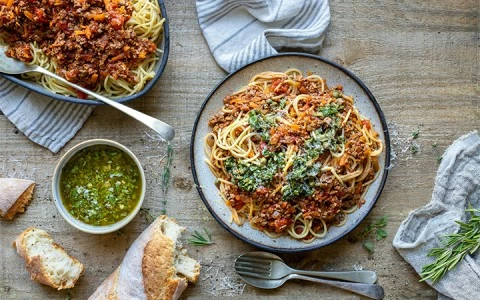 A plate of spaghetti with meat sauce and green herbs on top, accompanied by bread and a bowl of green sauce.