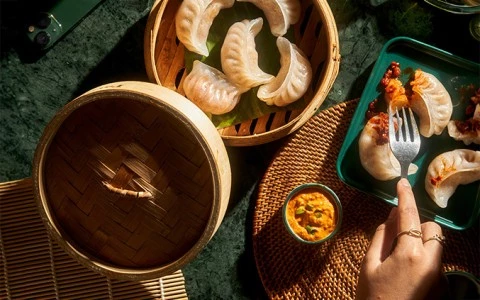A steamer basket of dumplings and a hand holding a fork with dumplings on a plate