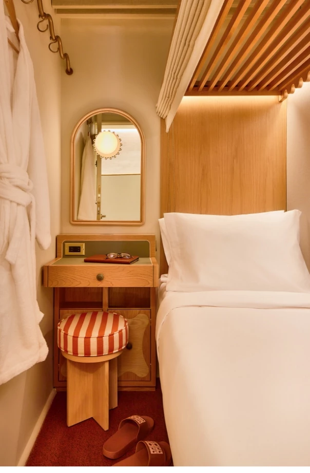 A small hotel room with a single bed, desk, mirror, and stool.