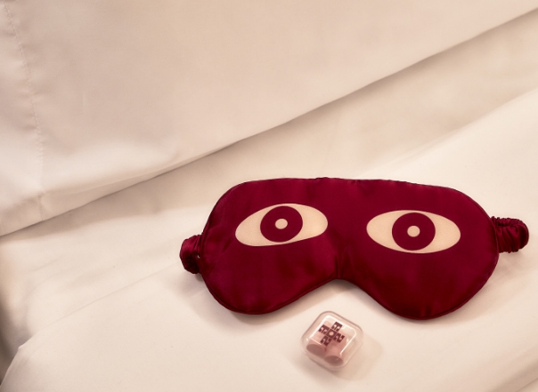 A red sleep mask with white eyes and a pink dice on a bed with white sheets.