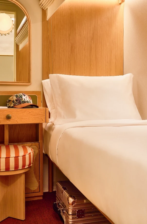 A close-up of a bed with white linens and a wooden headboard, with a floral hat on a nightstand and a suitcase underneath the bed.