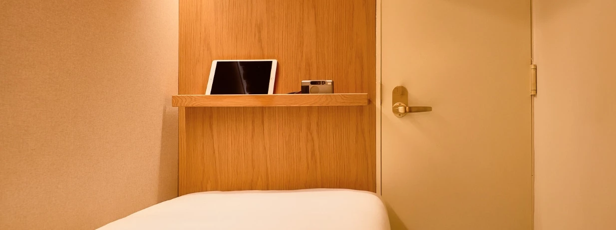 A small hotel room with a bed, a shelf with a tablet and a cup, and a door with a gold handle.