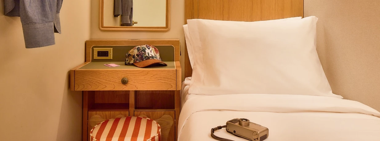 A close-up view of a bed with white linens and a wooden nightstand with a telephone, a hat, and a mirror on the wall.