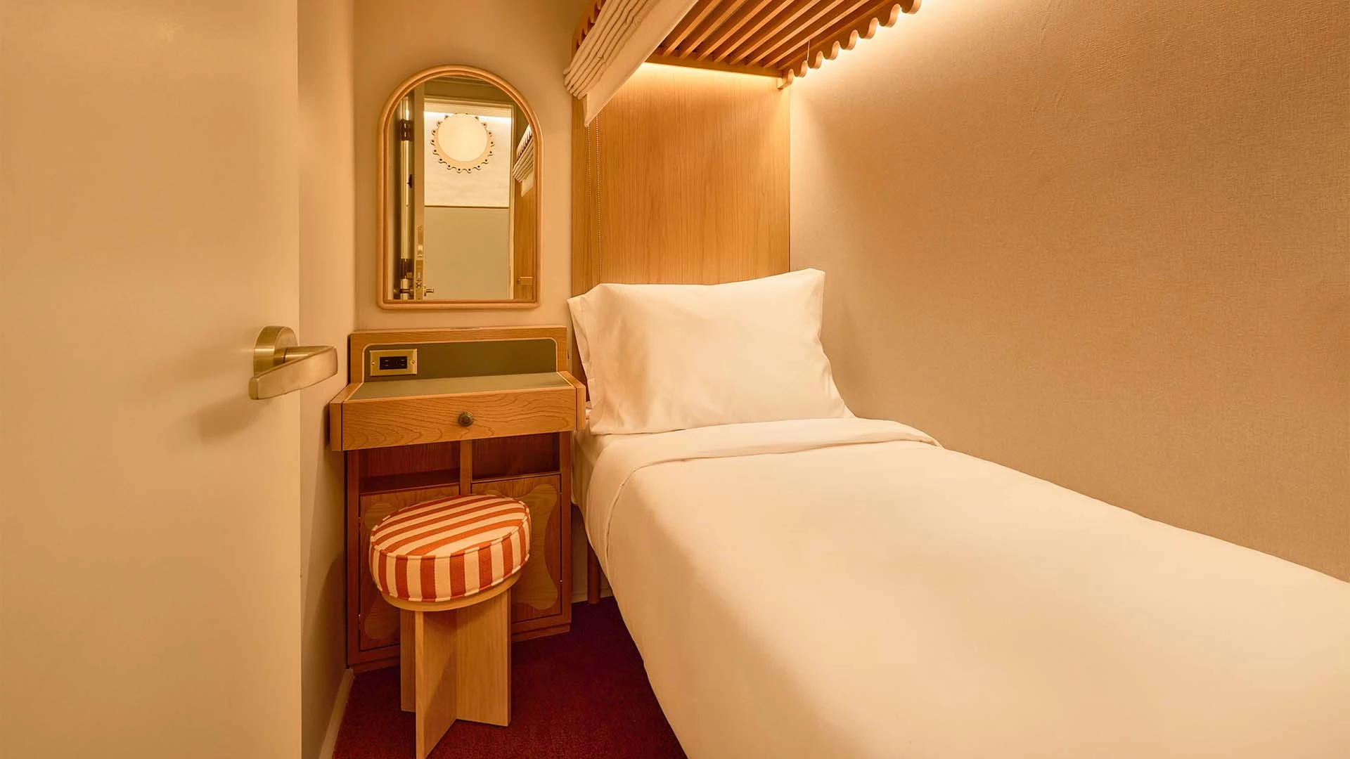 A small hotel room with a single bed, desk, mirror, and stool.