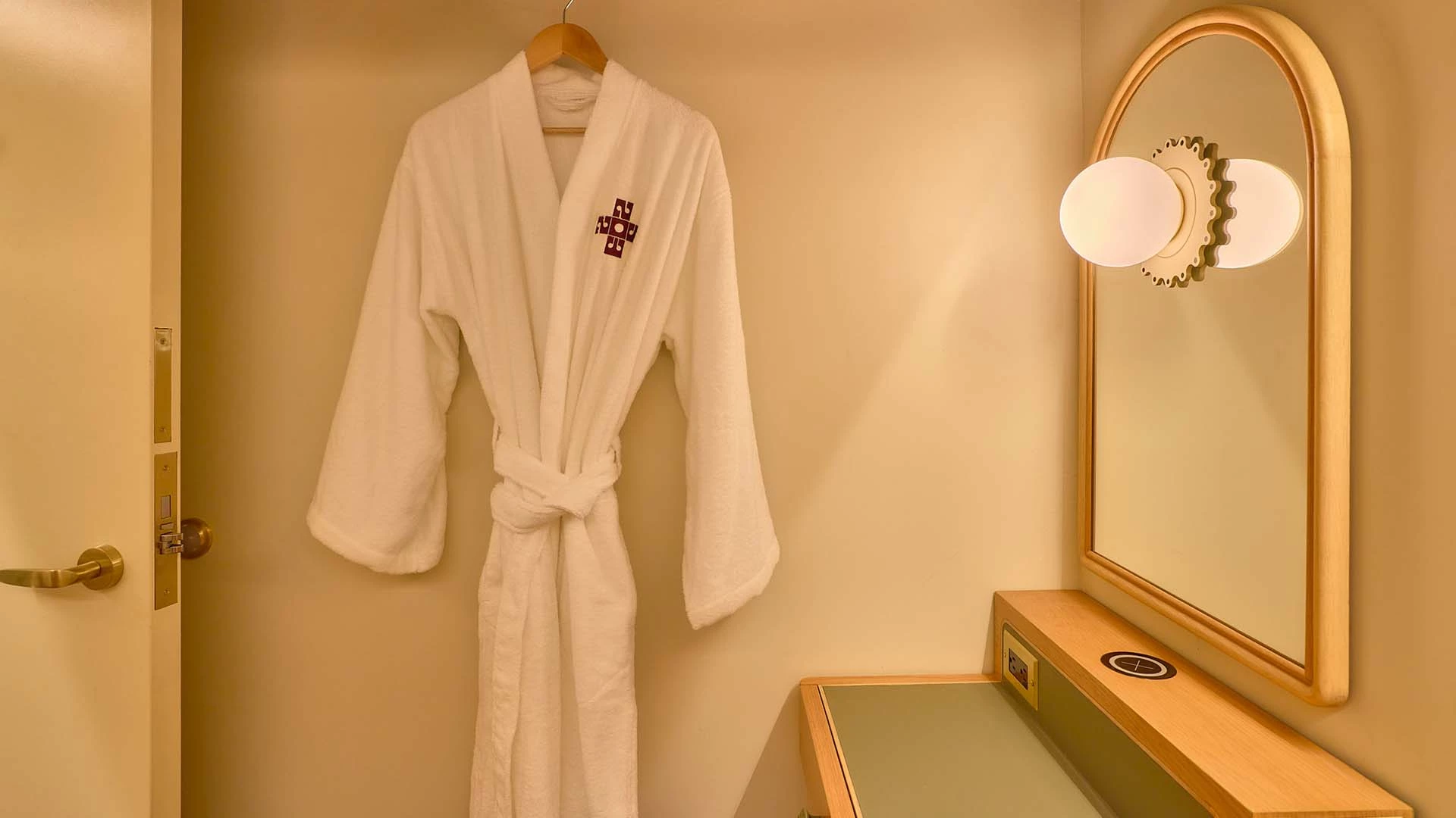 A white robe hangs on a hook next to a vanity with a mirror and lighting.