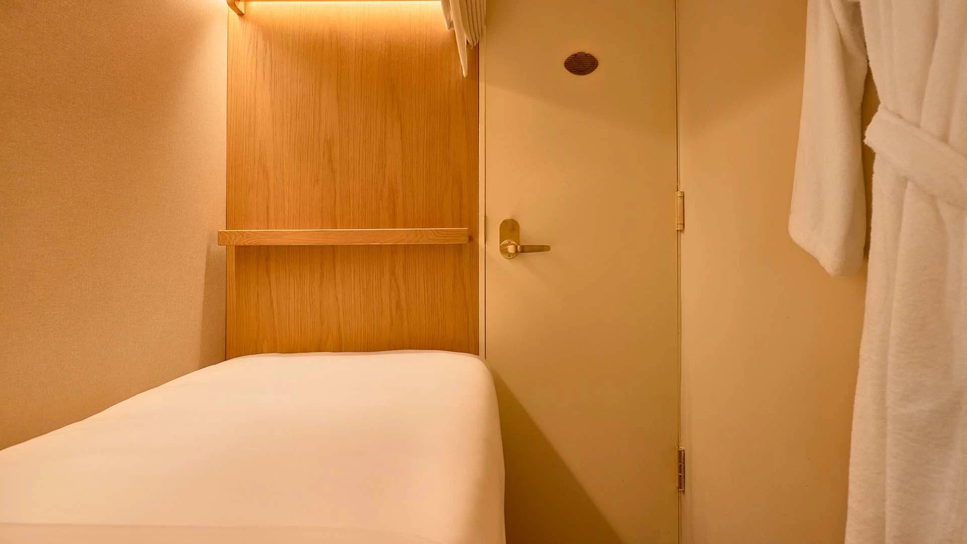 A small hotel room with a single bed, wood paneling, and a white robe hanging on the door.