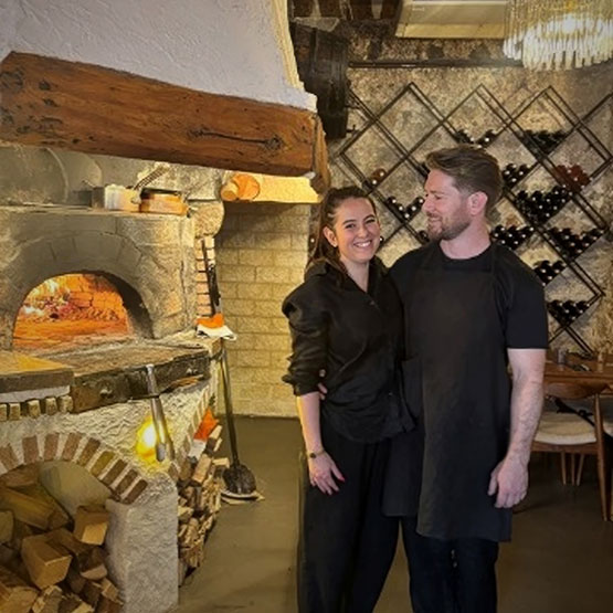 a man and woman standing in a room with a brick oven