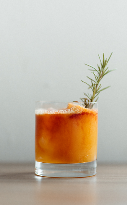 a glass of orange liquid with a sprig of rosemary