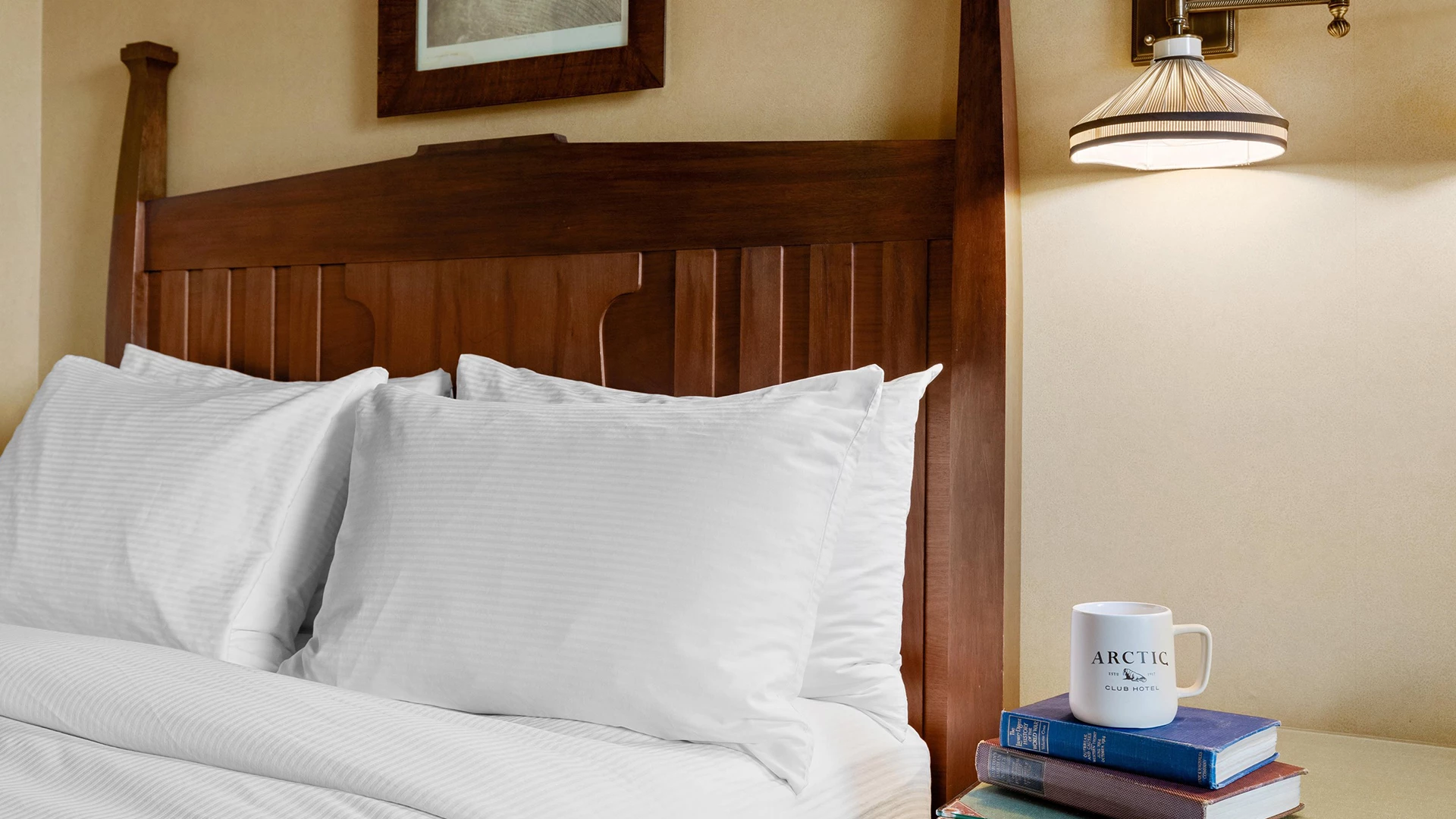 A close-up of a bed with a wooden headboard, white bedding, and a stack of books with a mug on top.
