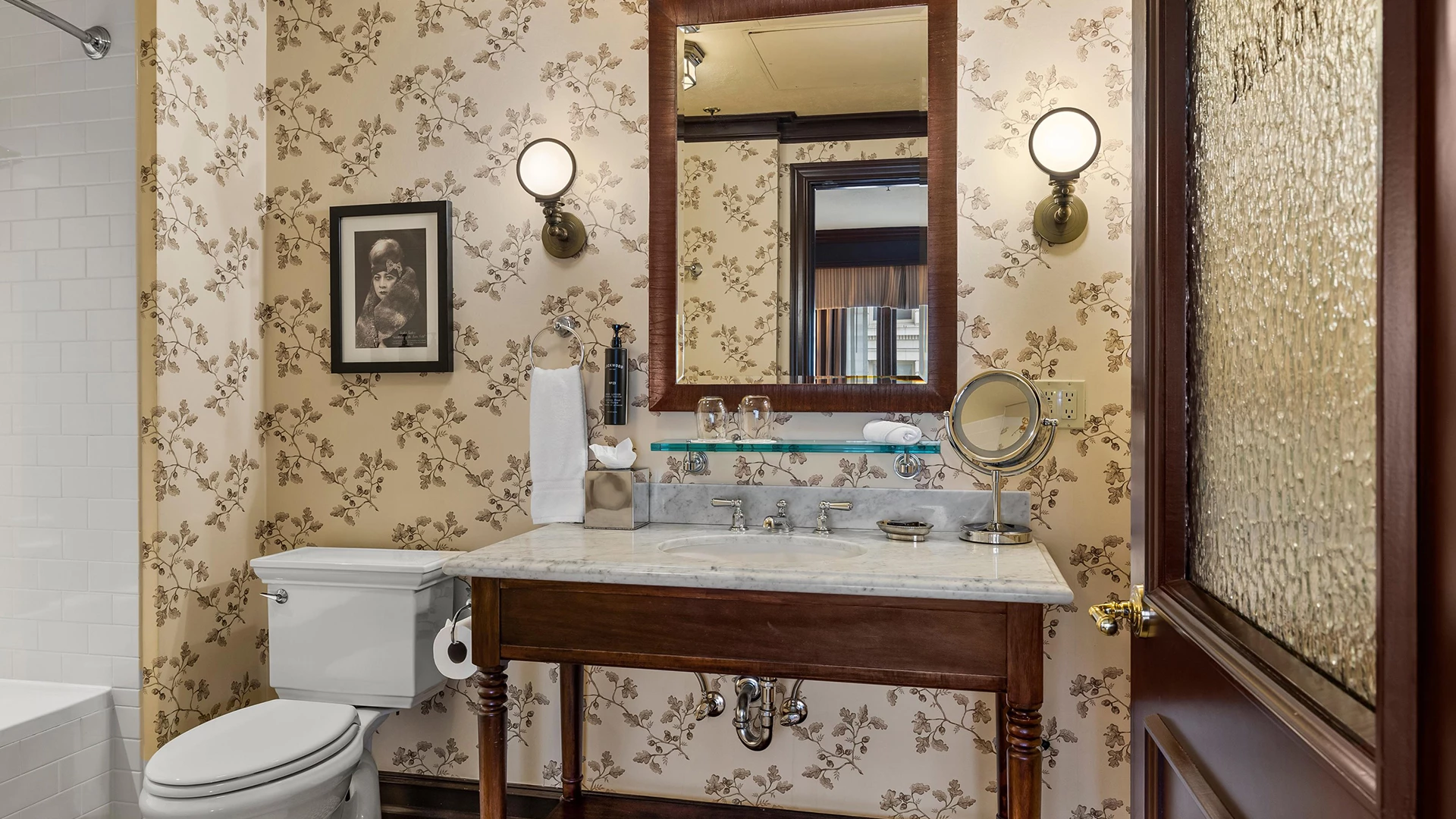 A bathroom with a marble countertop, wooden vanity, floral wallpaper, and a mirror above the sink.