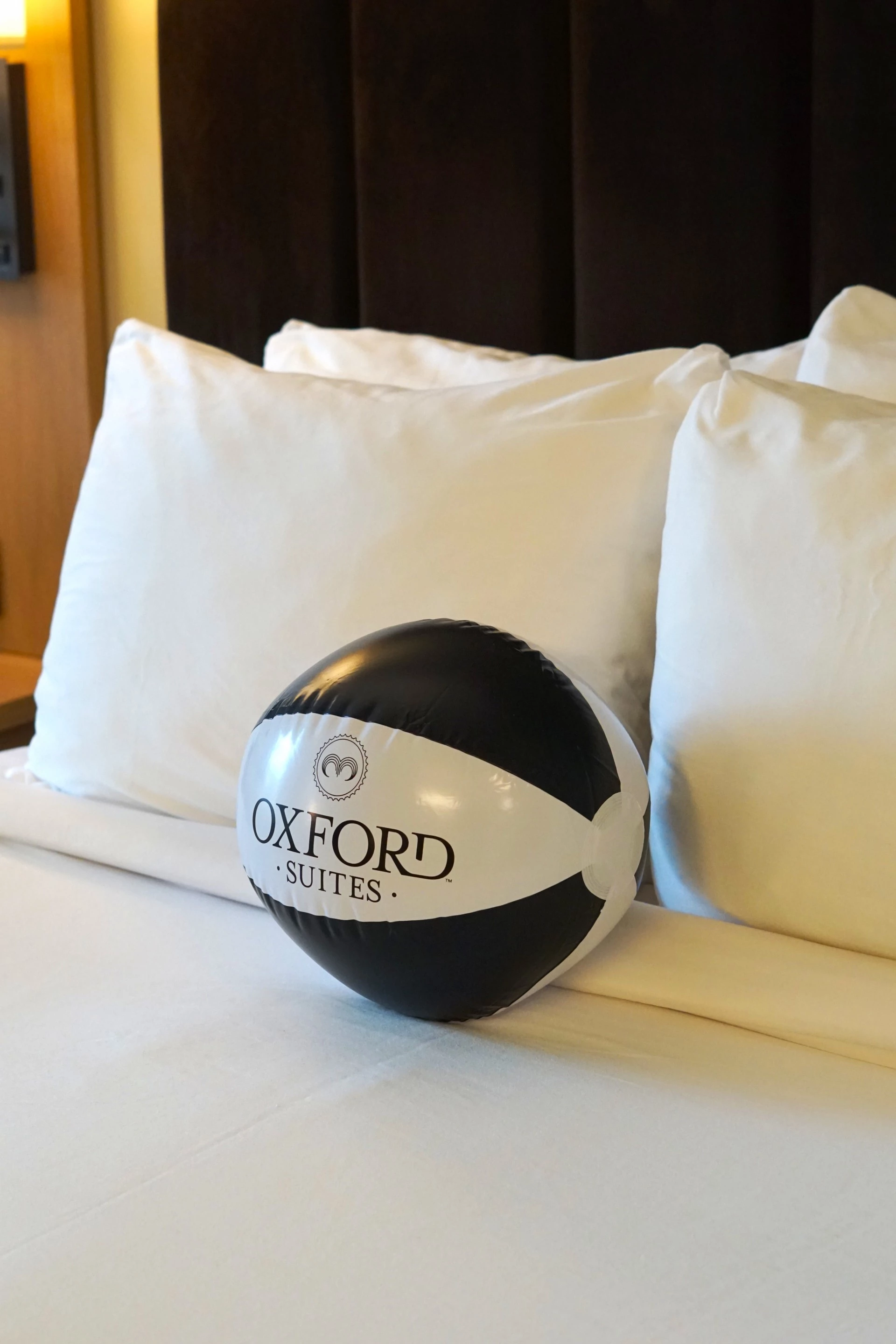 A black and white beach ball with 'OXFORD SUITES' printed on it sits on a bed with white pillows and sheets.