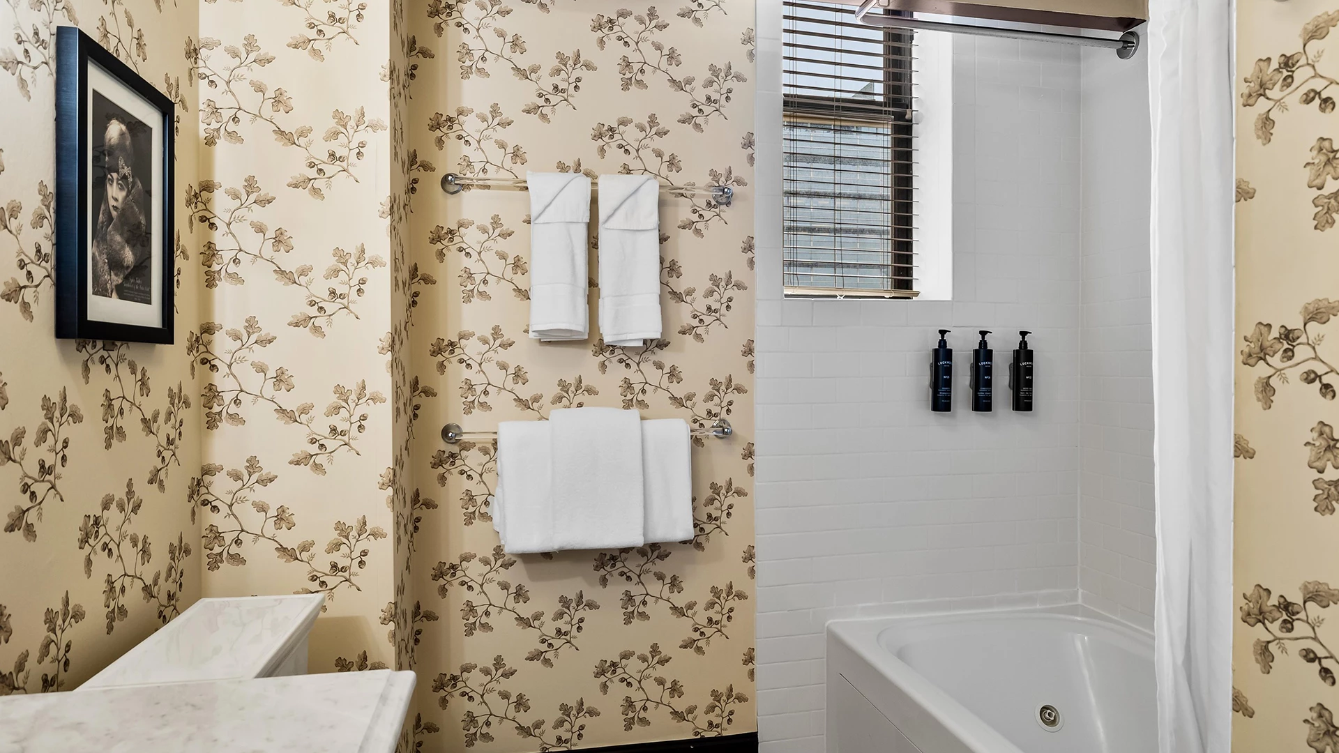 A bathroom with a tub, shower curtain, towels, toiletries, and floral wallpaper.