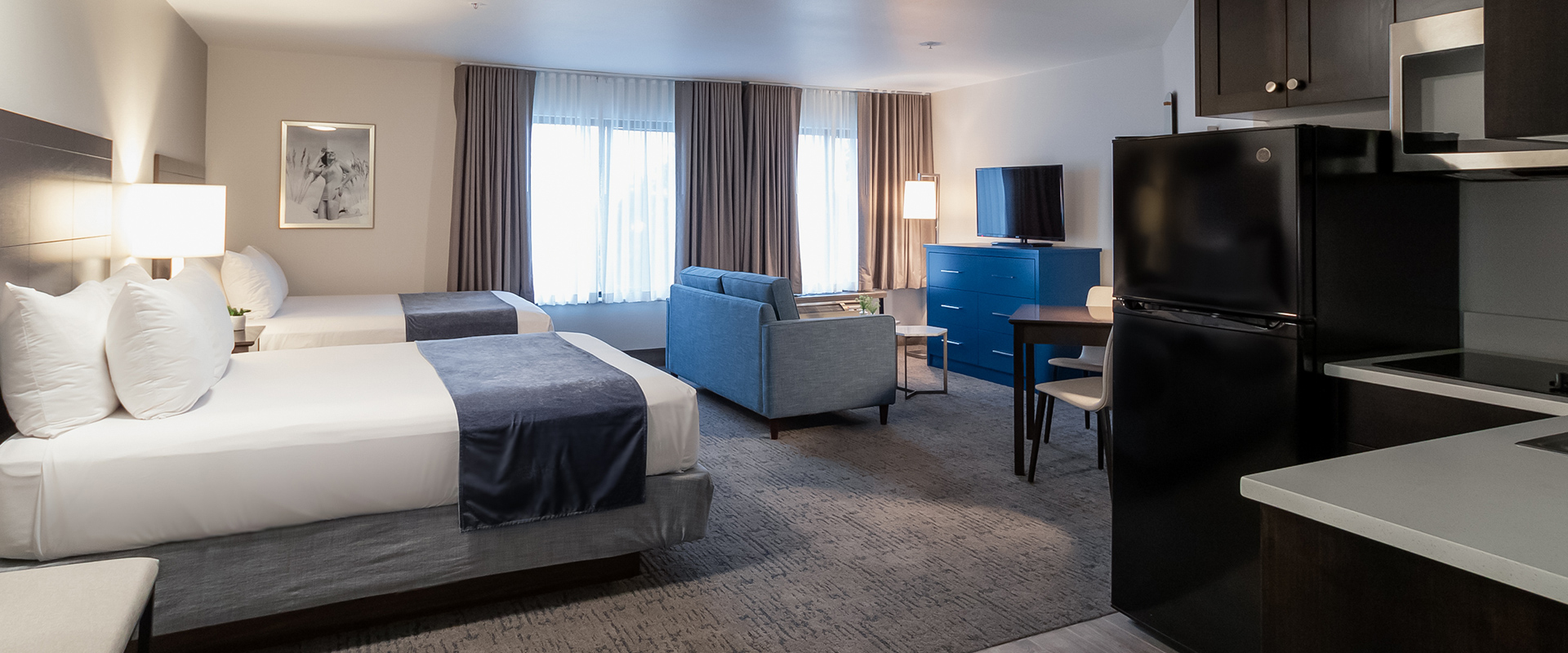 A hotel room with two beds, a sofa, a kitchenette, and a TV.