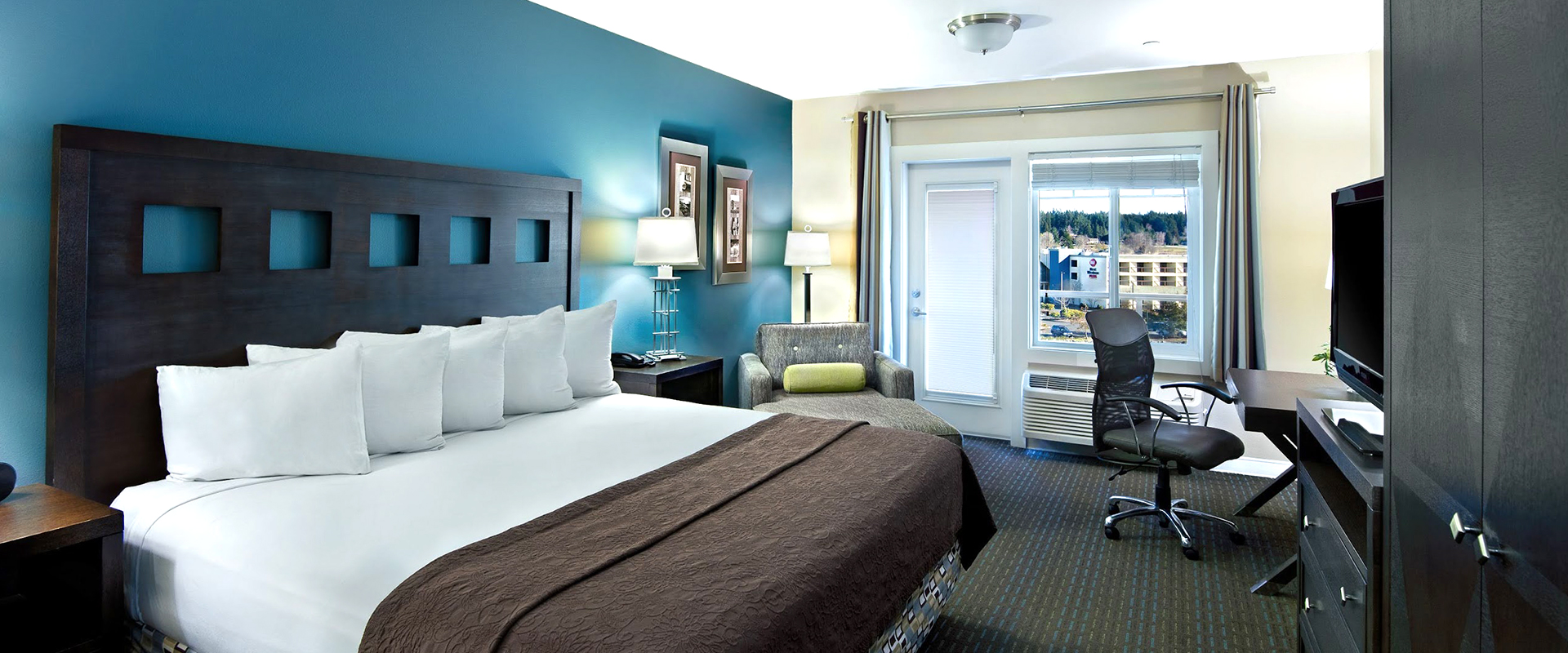 A hotel room with a large bed, desk, chair, and balcony door.