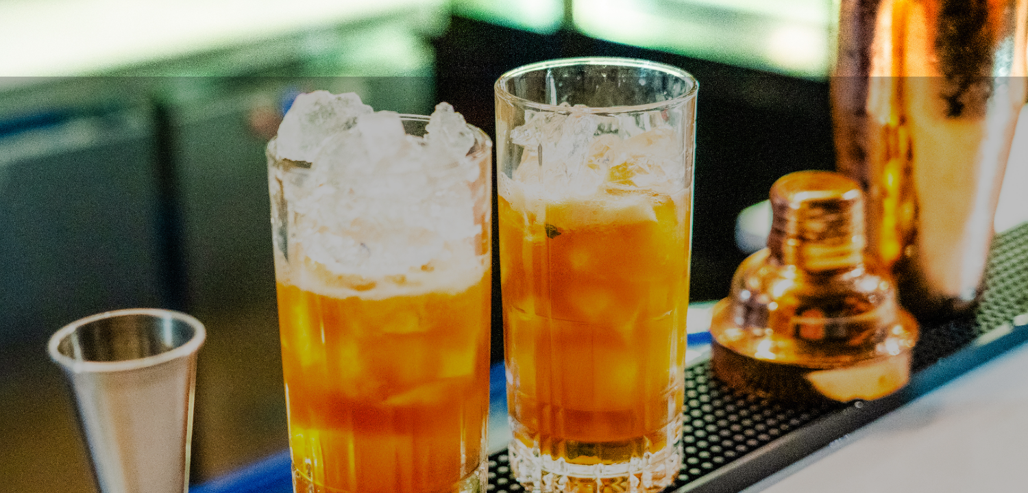 two glasses of liquid and ice on a bar