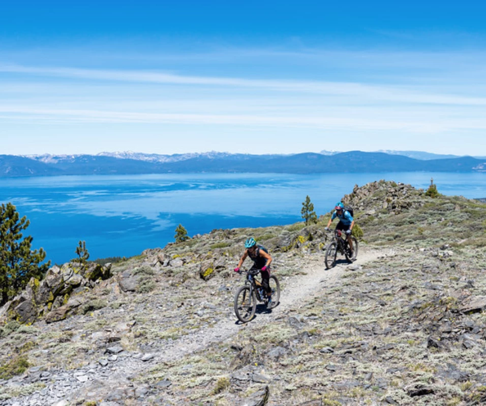 two people riding bikes on a rocky trail with a body of water in the background