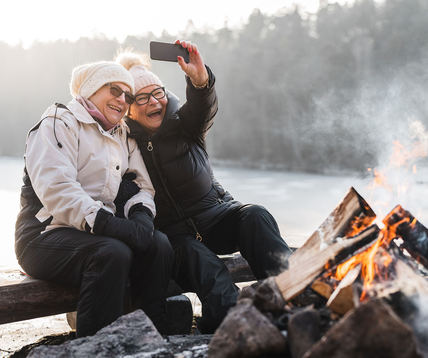 two women sitting on a log taking a selfie by a fire