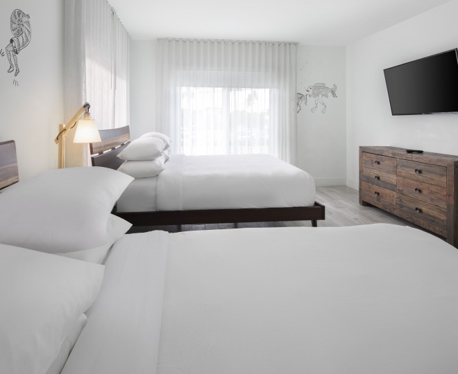 A bright and clean hotel room with two beds, a wooden dresser, and a flat-screen TV mounted on the wall.