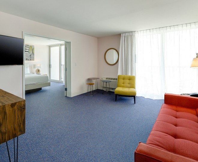 A wide view of a hotel room with a red couch, yellow chair, and doorway to a bedroom.