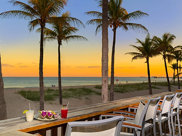 A beachside dining setup with a plate of food and drinks on a wooden table, overlooking the ocean at sunset.