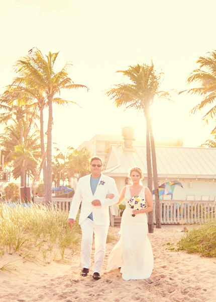 A couple in wedding attire walking on the beach with palm trees in the background.