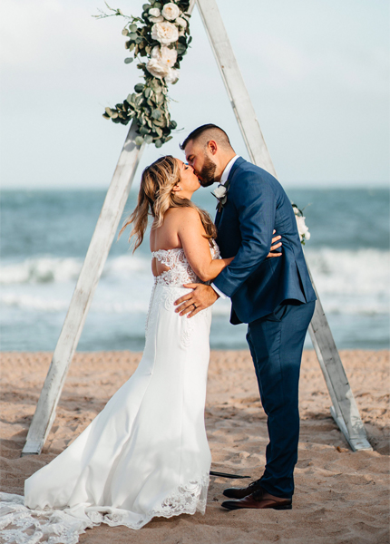 A couple in wedding attire embracing in front of a beach wedding arch.