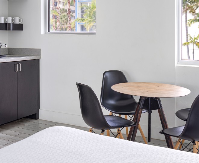 A modern hotel room with a kitchenette, dining table, and bed, showing a view of palm trees outside the window.