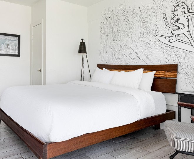 A neatly made bed with white linens and a wooden frame, accompanied by a chair and a lamp, in a room with a wall mural.