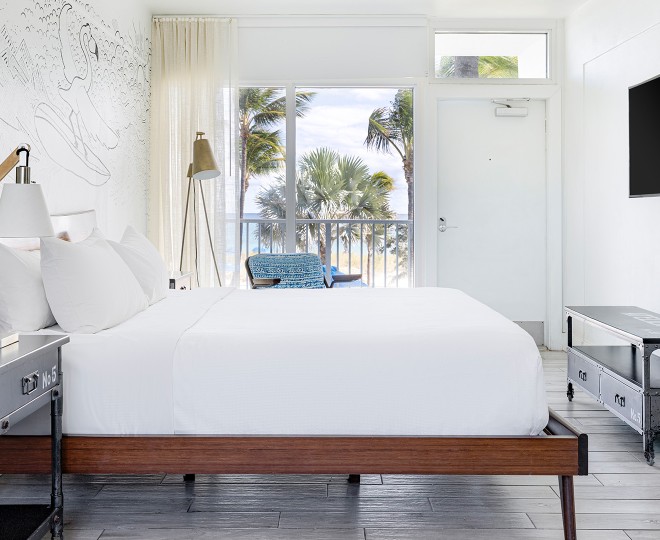 A white guest room with a large bed, wooden bed frame, and a large window looking out to palm trees.