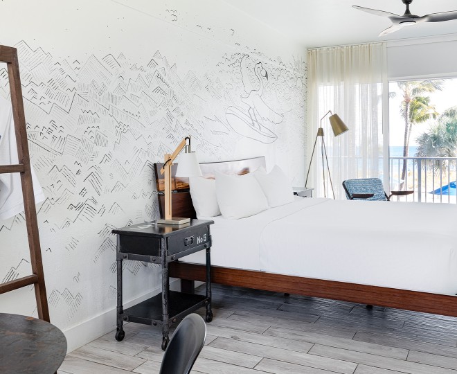 A white guest room with a large bed, nightstand, and window with a balcony.