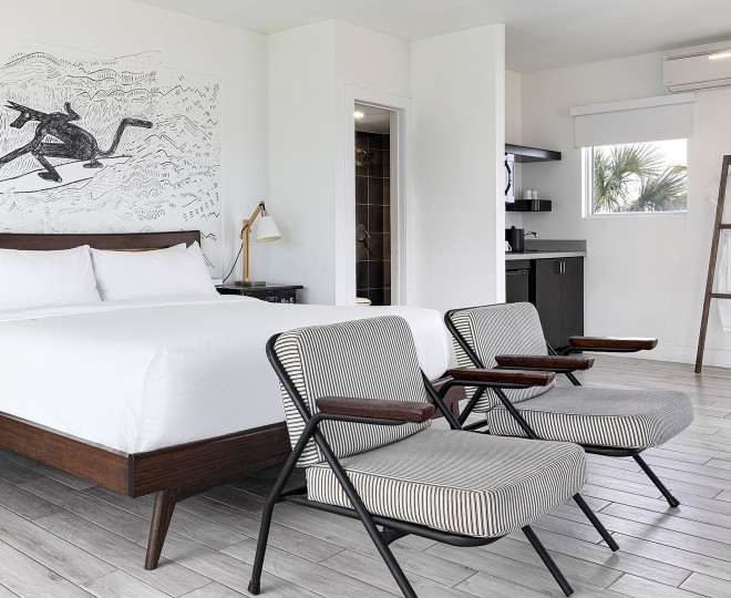 A wide view of a hotel room with a bed, two chairs, a kitchenette, and artwork on the wall.