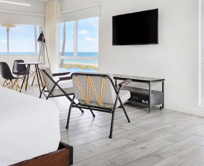 A bright and airy hotel room with a beach view, featuring a bed, dining table, chairs, and TV.