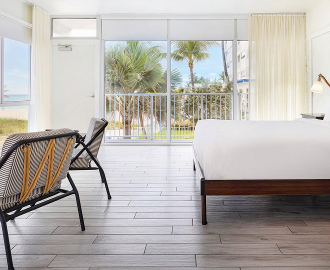 A bright, beach-style room with a large bed, chair, and sliding glass doors leading to a balcony with a view of palm trees and the ocean.