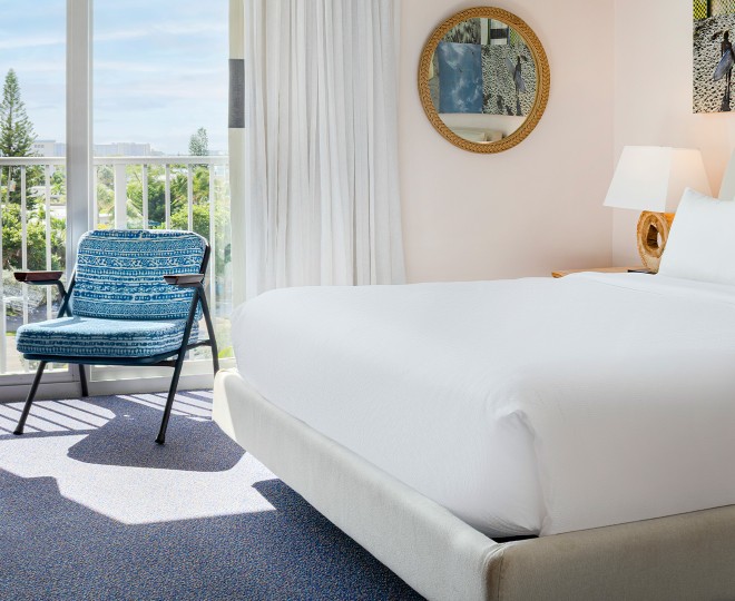 A neatly made bed with white bedding and a blue chair in front of a curtained window.