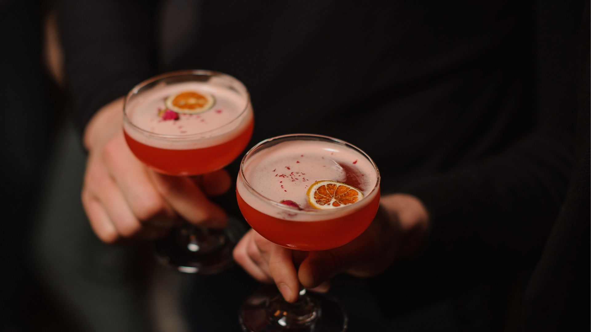 a pair of hands holding glasses with pink liquid and orange slices