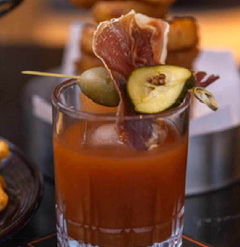 a glass of liquid with a pickle and a slice of meat on top die hard
