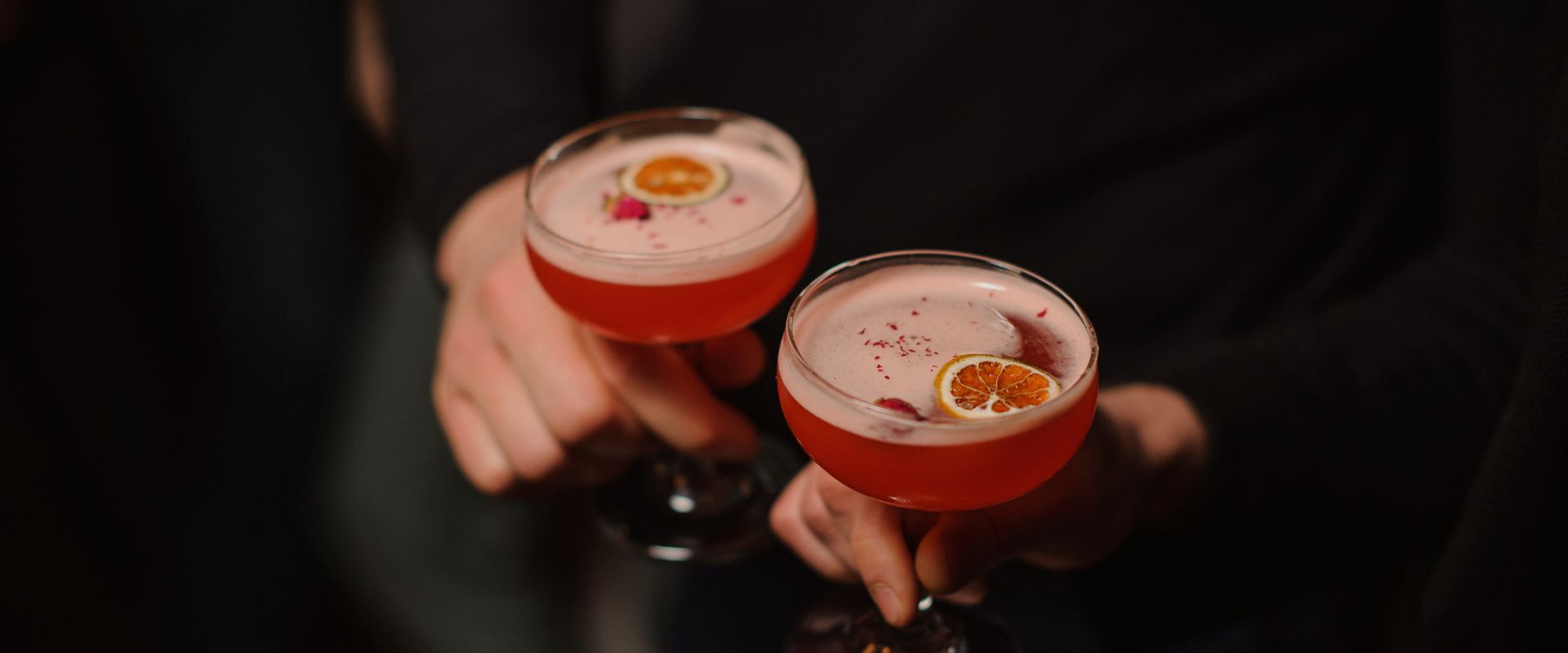 a pair of hands holding glasses with pink liquid and orange slices