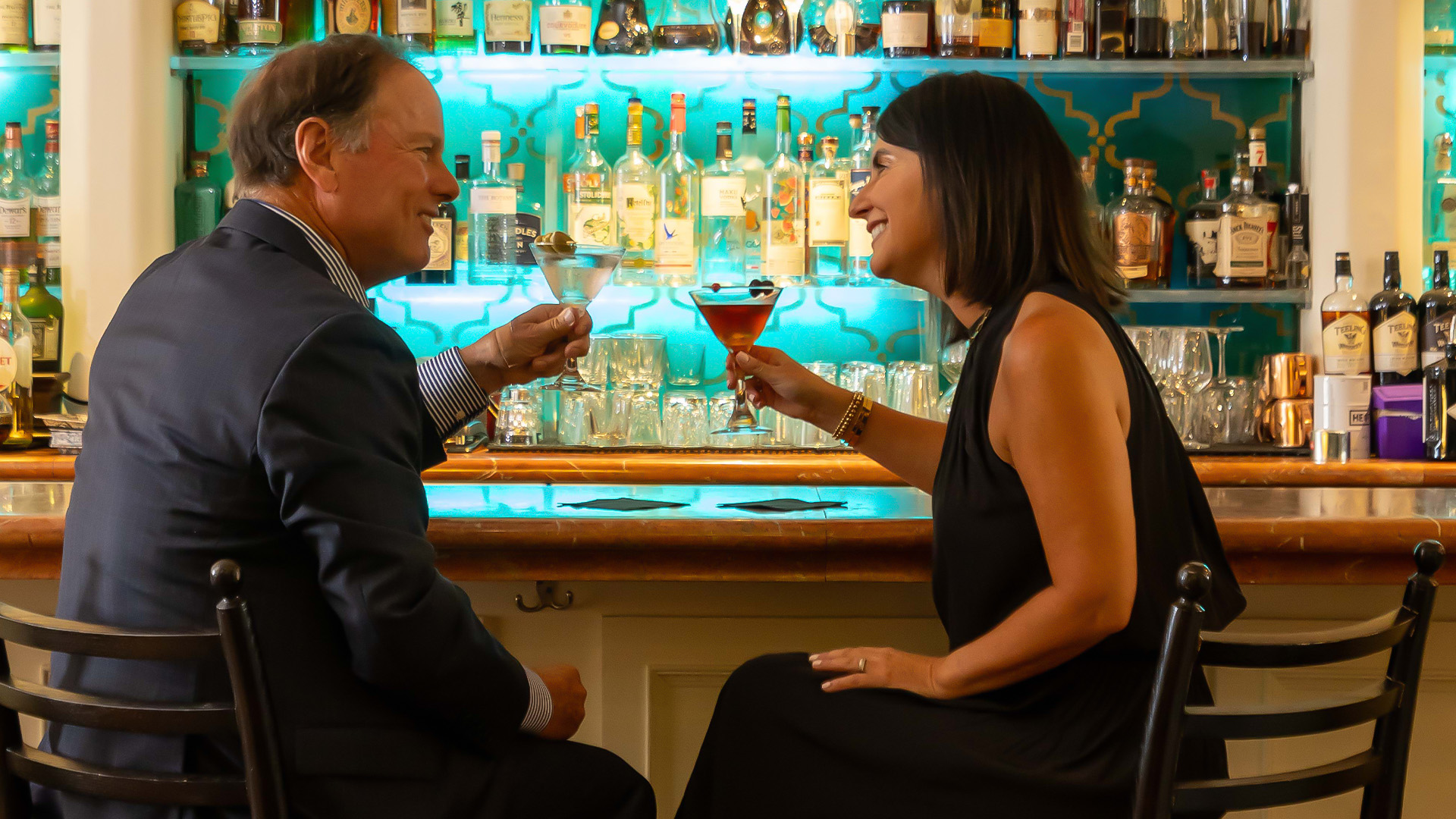 a man and woman sitting at a bar toasting with drinks