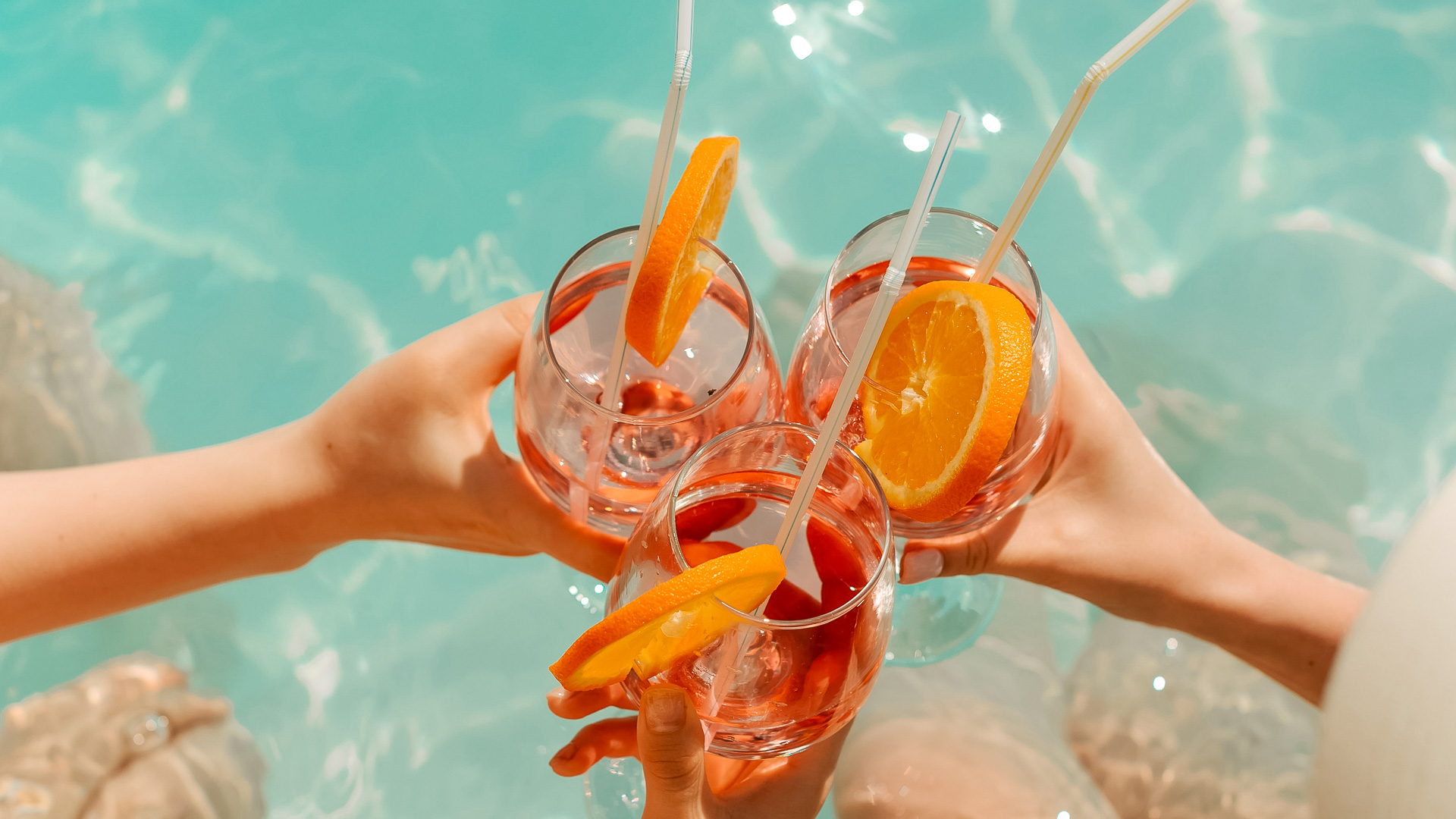 a group of hands holding glasses with orange slices and straws in front of water