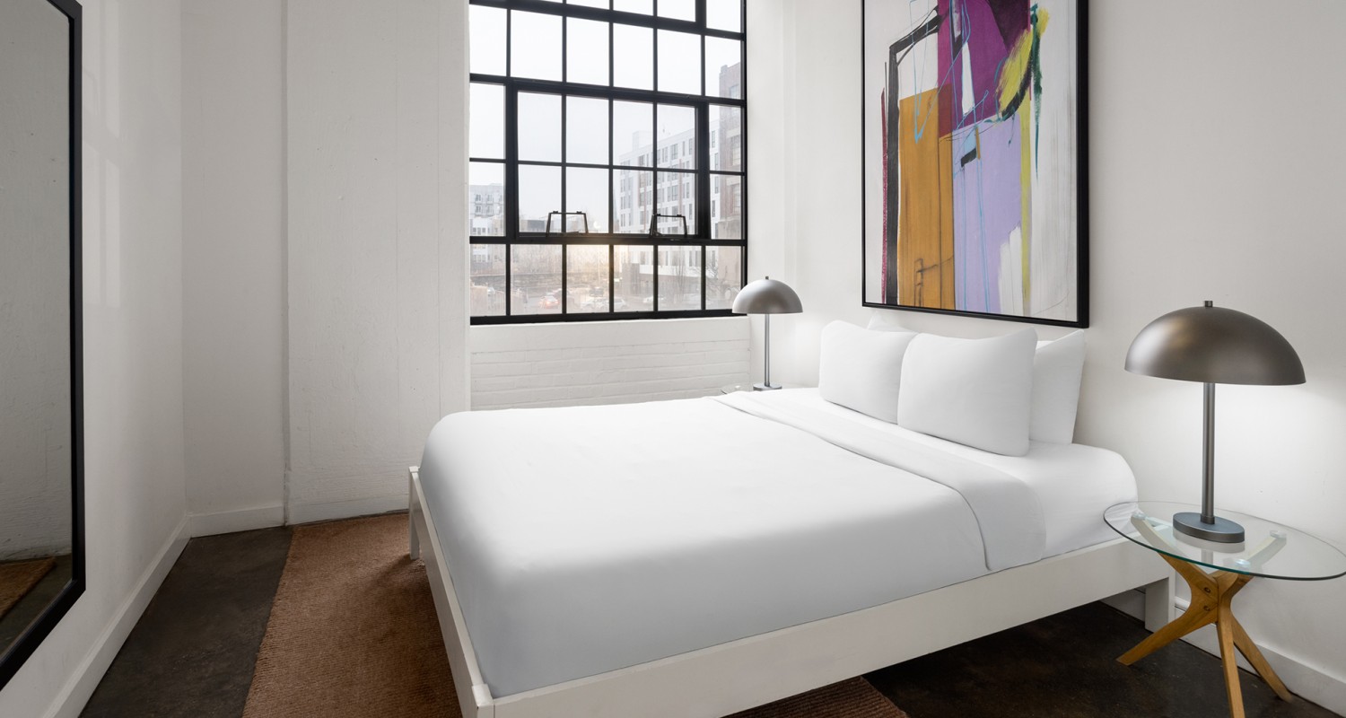 a bed with white sheets and a lamp in a room with large windows