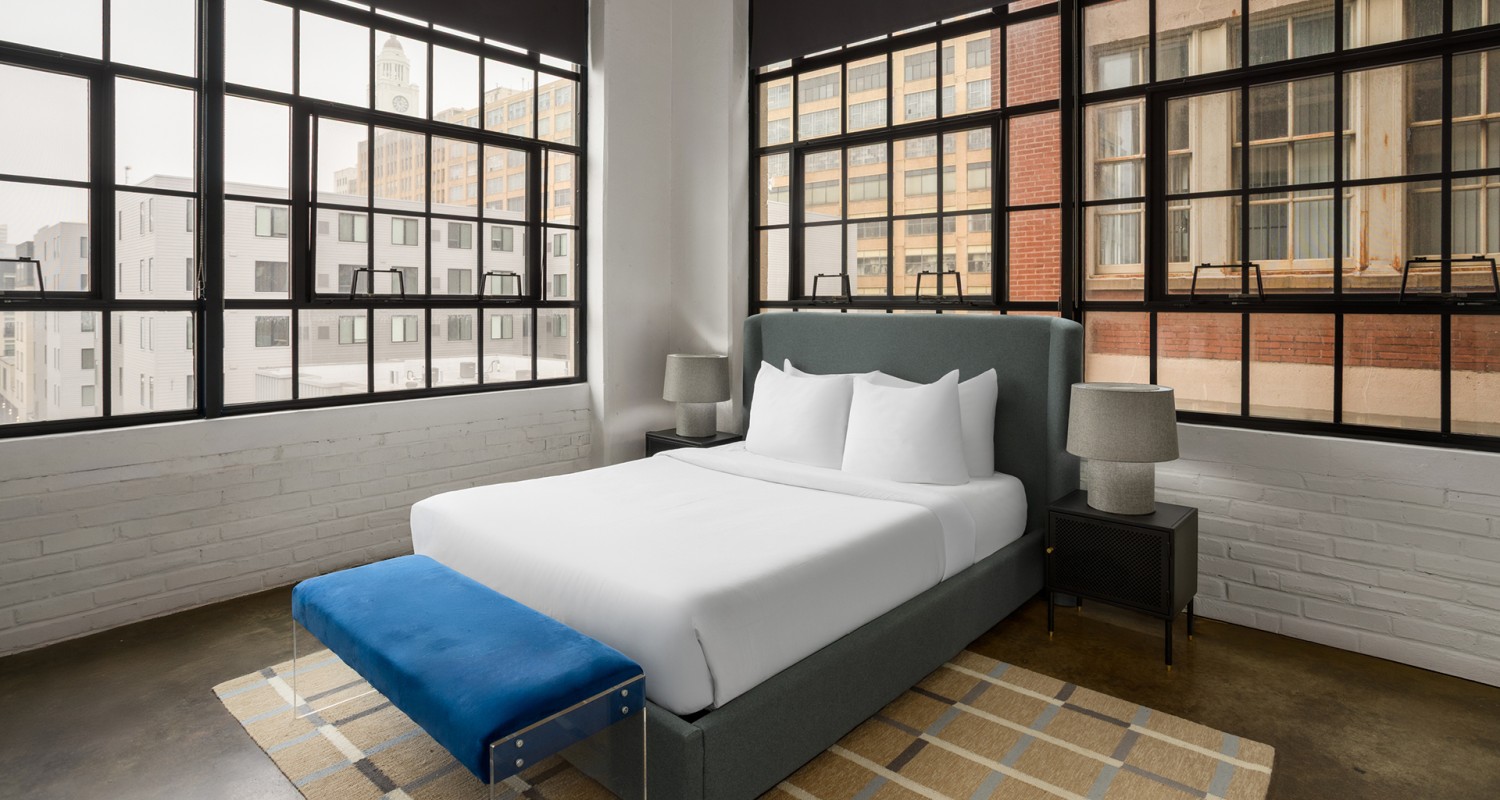 a bed with white sheets and a blue bench in a room with windows