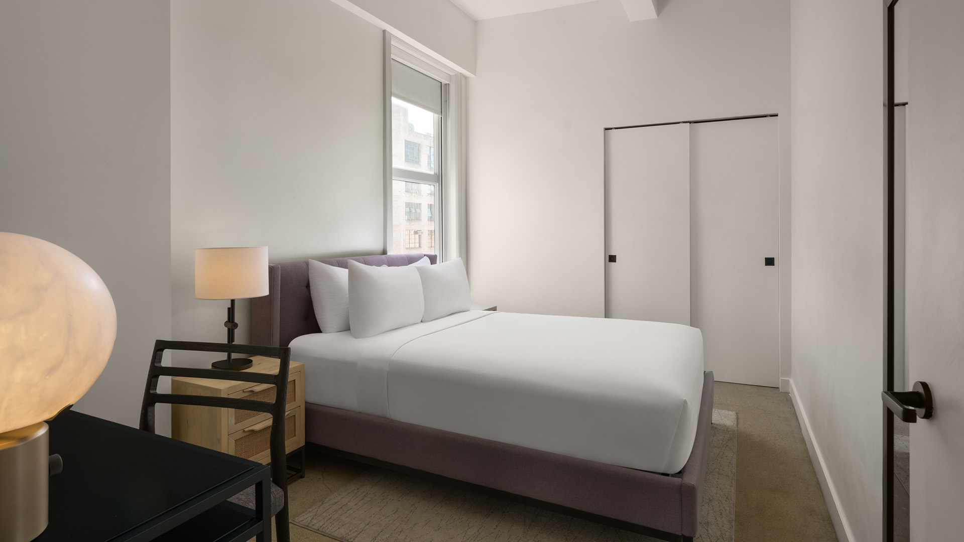 A modern, minimalist hotel bedroom with a plush bed topped with crisp white linens, a soft purple upholstered bed frame, a wooden nightstand with lamp, and sliding closet doors.