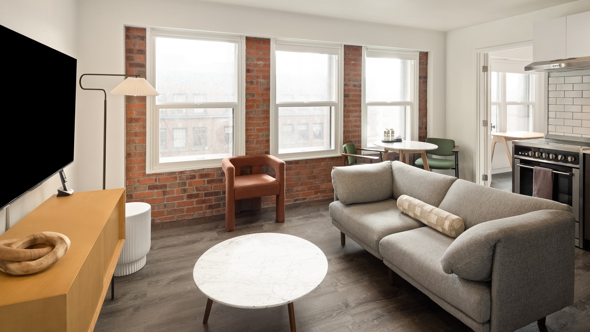 A modern apartment living room featuring exposed red brick walls with large windows, a grey fabric sofa with a cylindrical bolster pillow, a round marble coffee table, a rust-orange accent chair, and a glance into an adjoining kitchen area.