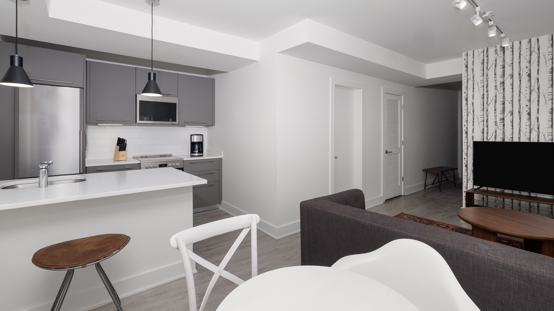 A modern open-plan hotel suite featuring a kitchen counter with bar seating, matte grey cabinetry, a living area with a dark grey sofa, and a television against birch tree wallpaper.
