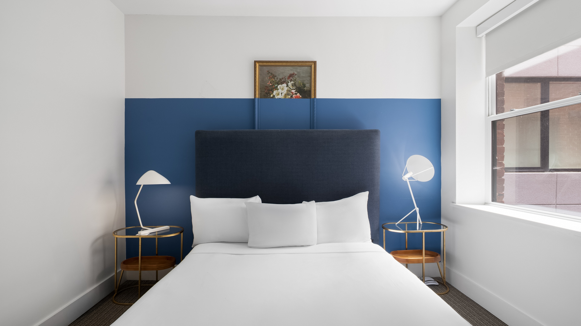 A stylish guest room featuring a bed with a dark navy padded headboard, crisp white linens, a blue color-blocked accent wall, round brass nightstands, and modern white reading lamps.