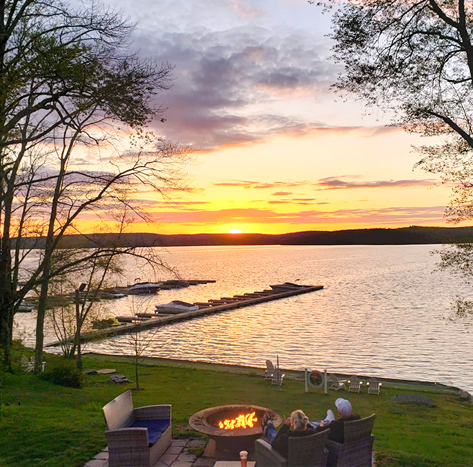 a group of people sitting on a patio by a fire pit with a lake and dock