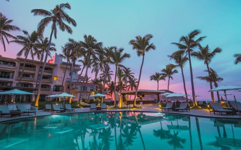 a pool with palm trees and a building with a pink sunset