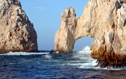 a large rock formation with a arch in the middle of the ocean with Arch of Cabo San Lucas in the background